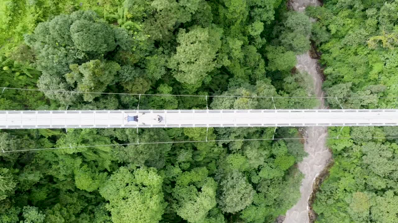 Stunning top-down aerial footage of a pedestrian suspension bridge spanning a river in the Himalayas. Captures the dramatic view of the bridge amidst rugged terrain and rushing waters.