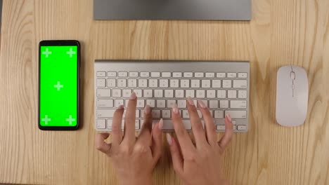 businesswoman is typing on the keyboard in the office or at home. smartphone with green screen and chroma key with vertical screen position. desktop top view. hands close up. slow motion