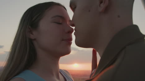 close up romantic couple standing in hot air balloon basket overlooking golden sunset horizon as faces glow in warm light while paraglider drifts in distance above mosaic fields at tranquil dusk