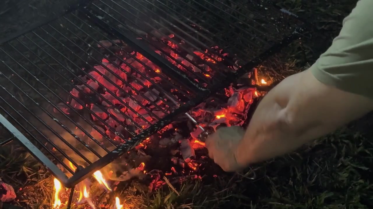 Argentine asado grill positioned over glowing embers and open fire, with smoke rising as hot coals are moved and redistributed beneath the cooking grate