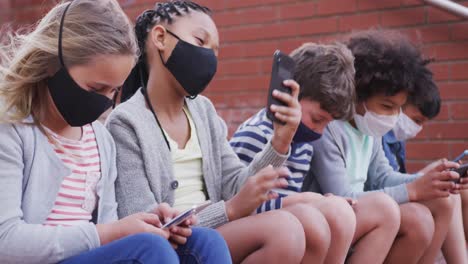 group of kids wearing face masks using smartphones while sitting together