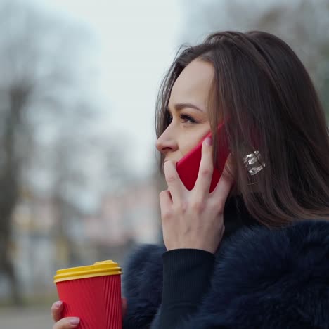 Attractive young girl in fur coat. Beautiful woman in stylish coat talking the phone and holds coffee glass in the park.