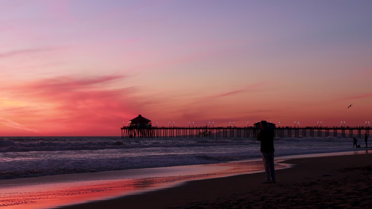 A man takes a picture of the beach during a gorgeous red, purple, tangerine, pink and blue sunset with the Huntington Beach Pier in the background at Surf City USA California