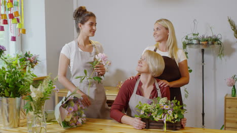 titolare di un negozio di fiori di mezza età seduto al tavolo di legno con squisita esposizione floreale e in posa con due giovani fioristi, con mazzi di fiori e bouquet sullo sfondo