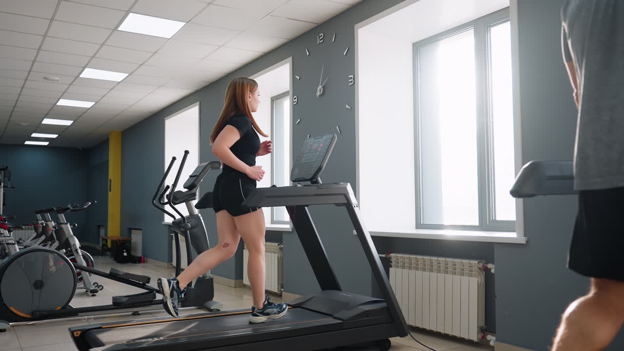 side view of athletic woman running at high speed on treadmill in bright gym interior with sunlight streaming through large windows and partial view of man walking slowly on adjacent machine
