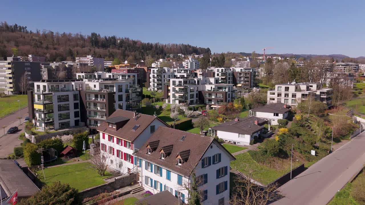 Aerial rising shot of modern and luxury residential apartments with large windows in Switzerland. Wide shot. Forest trees in mountain in distance Sunny sporing day in small city. Upper class houses.