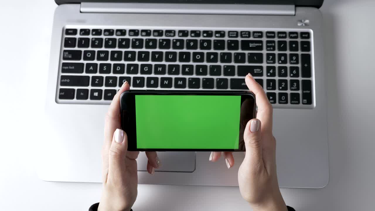 Female hands holding the smartphone. Green screen, chromakey concept, laptop in the background. Top shot 60 fps