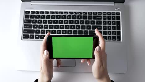 female hands holding the smartphone. green screen, chromakey concept, laptop in the background. top shot 60 fps