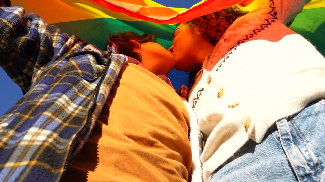 Low angle view of a lesbian couple kissing under a large rainbow flag on a sunny day