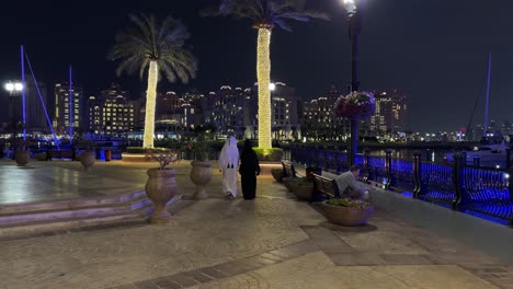 Pareja-árabe-Hombre-Mujer-Con-Un-Vestido-De-Moda-Local-Tradicional-Bisht-Hijab-Caminando-Playa-De-Lujo-En-Katara-Pearl-Qanat-Quartier-En-Doha-Qatar-El-Diseño-Del-Paisaje-De-La-Ciudad-Está-Decorado-Con-Palmeras-Y-Luz