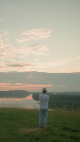 un caballero está contemplativo en un campo cubierto de hierba junto a un lago tranquilo al atardecer. vestido con una camisa blanca, sombrero y vaqueros, captura un momento de paz y reflexión mientras el sol se pone