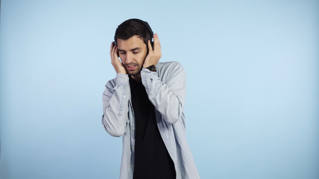 hombre caucásico satisfecho poniendo sus auriculares negros y comienza a bailar al ritmo de la melodía, cierra los ojos escucha una canción fuerte en los auriculares sobre fondo azul