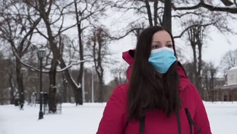 mujer turista con abrigo encapuchado con máscara facial caminando en un parque de invierno nevado