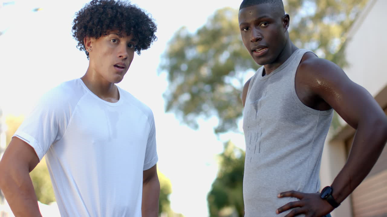 Exercising outdoors, two multiracial male friends taking break and looking at camera