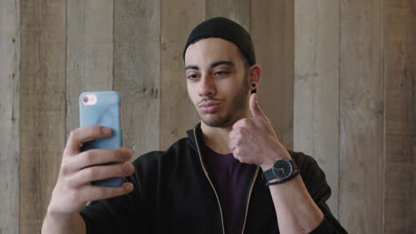 Primer-Plano-Retrato-De-Un-Atractivo-Joven-Estudiante-Hispano-Posando-Con-El-Pulgar-Hacia-Arriba-Gesto-Tomando-Una-Foto-Selfie-Usando-La-Tecnología-De-La-Cámara-Del-Teléfono-Inteligente