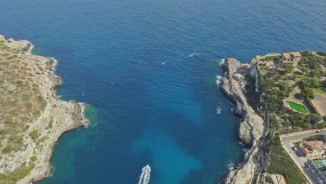 a dramatic aerial view of a coastal bay framed by rugged cliffs and deep blue water, with a speedboat leaving a white trail in its wake.
