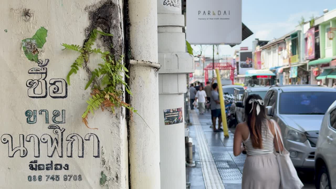 Woman strolls past street art and parked cars in vibrant, sunlit old town Phuket, Thailand