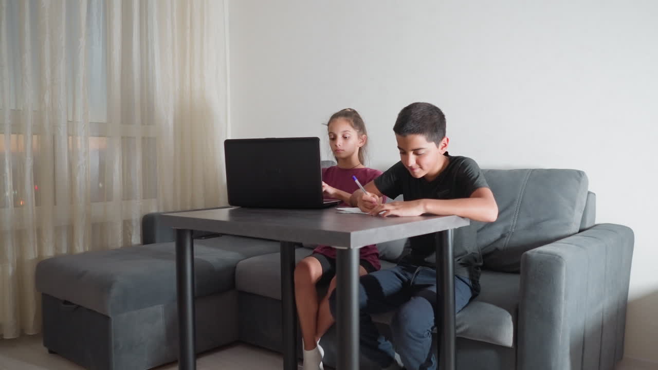 Hermanos haciendo los deberes sentados en el sofá del salón: un niño escribe en un papel y una niña teclea en un portátil, concentrados durante la sesión de estudio vespertina.