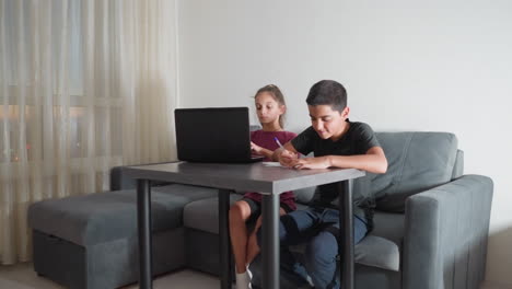 siblings doing homework seated on couch in living room with boy writing on paper and girl typing on laptop while concentrating during evening study session