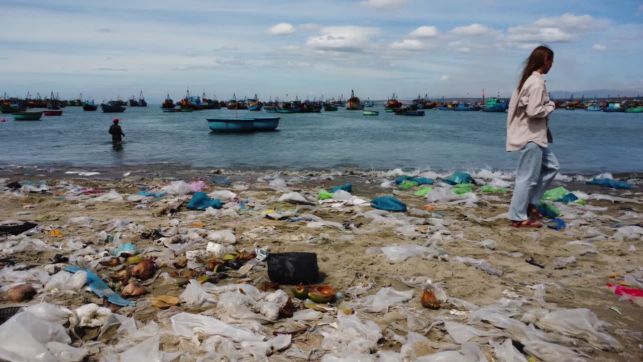 쓰레기 더미로 뒤덮인 모래 해변을 걷고 있는 슬픈 젊은 여성, 팬 오른쪽 보기