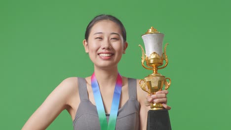 close up of asian woman with a gold medal and trophy smiling to camera with one arm akimbo posing as the first winner on green screen background in the studio