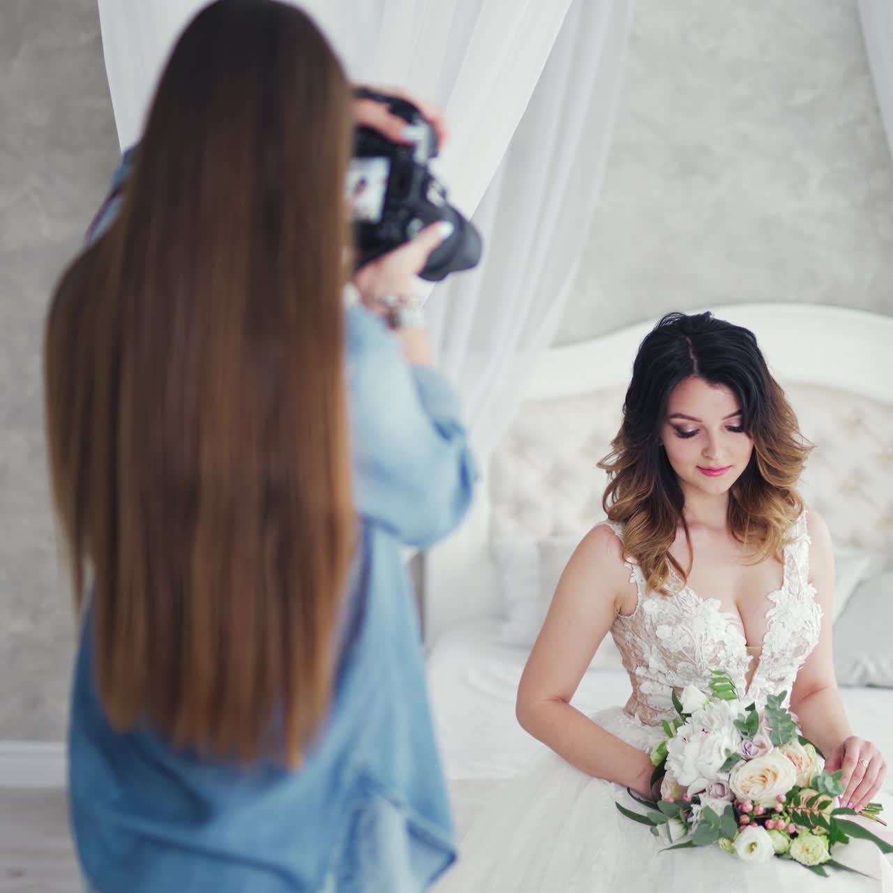 Female photographer with long dark hair is shooting by camera a pretty bride. Attractive young bride with flowers posing to the professional photographer on a white bed in studio.