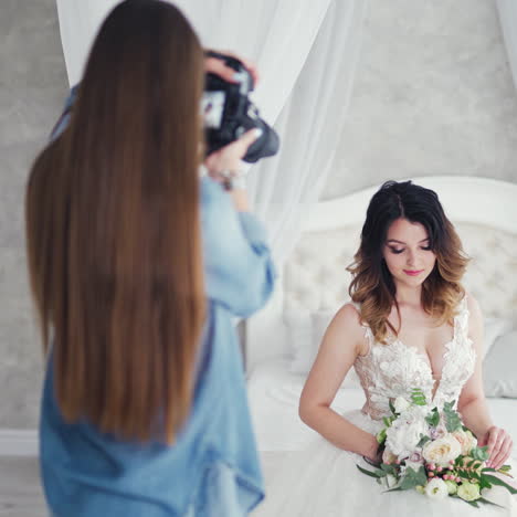 female photographer with long dark hair is shooting by camera a pretty bride. attractive young bride with flowers posing to the professional photographer on a white bed in studio.