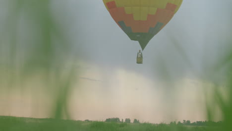 close up tall green grass framing hot air balloon carrying people and pilot firing burner flame from wicker basket, floating low above lush open field under gentle pastel sky