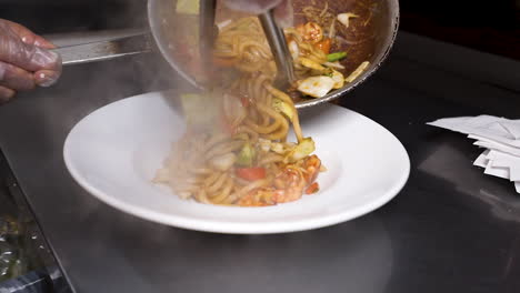 chef uses tongs to plate steaming hot freshly stir fried udon noodles on white plate, slow motion hd