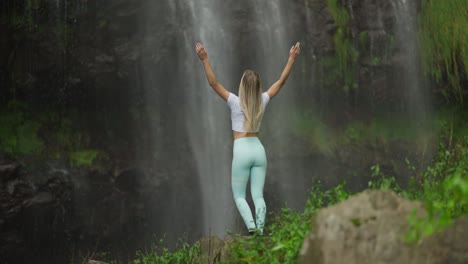 mujer en forma con ropa deportiva alcanzando la base de una cascada tropical levantando las manos