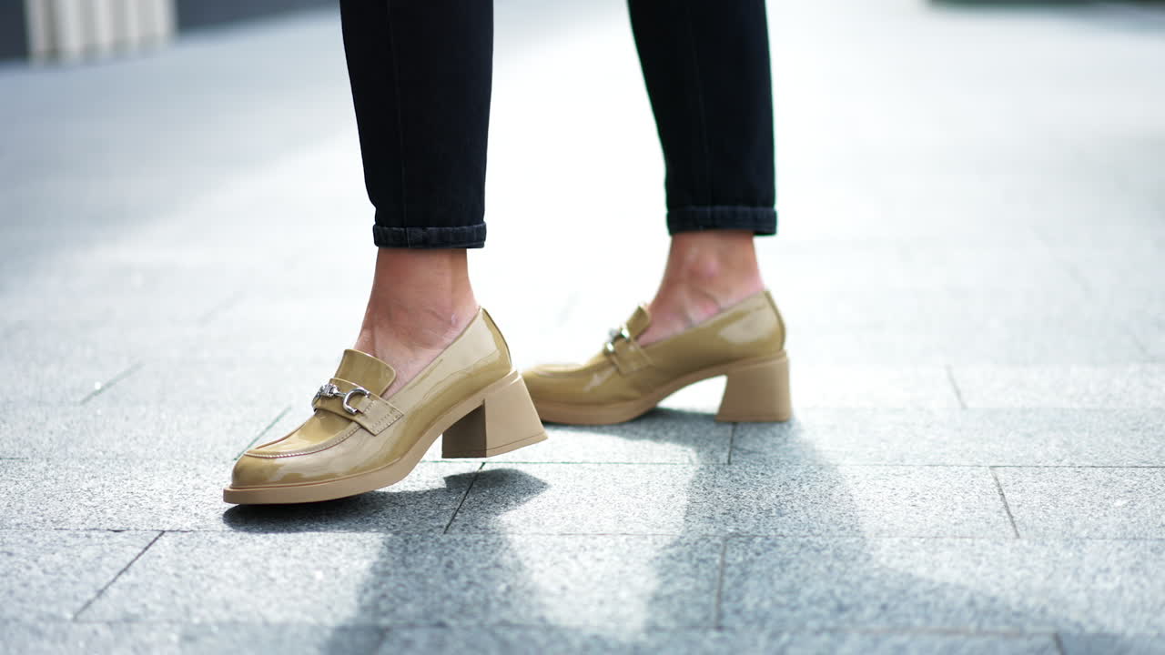 Beige patent leather shoes with metal decoration. Female model in black pants demonstrating the footwear. Close up.