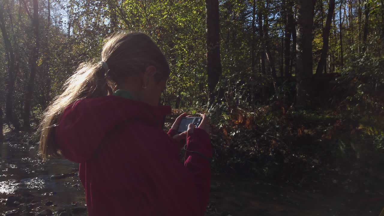 Sunlight filters through trees as a young girl with a ponytail, wearing a maroon jacket, stands by a river and takes photos with a digital camera
