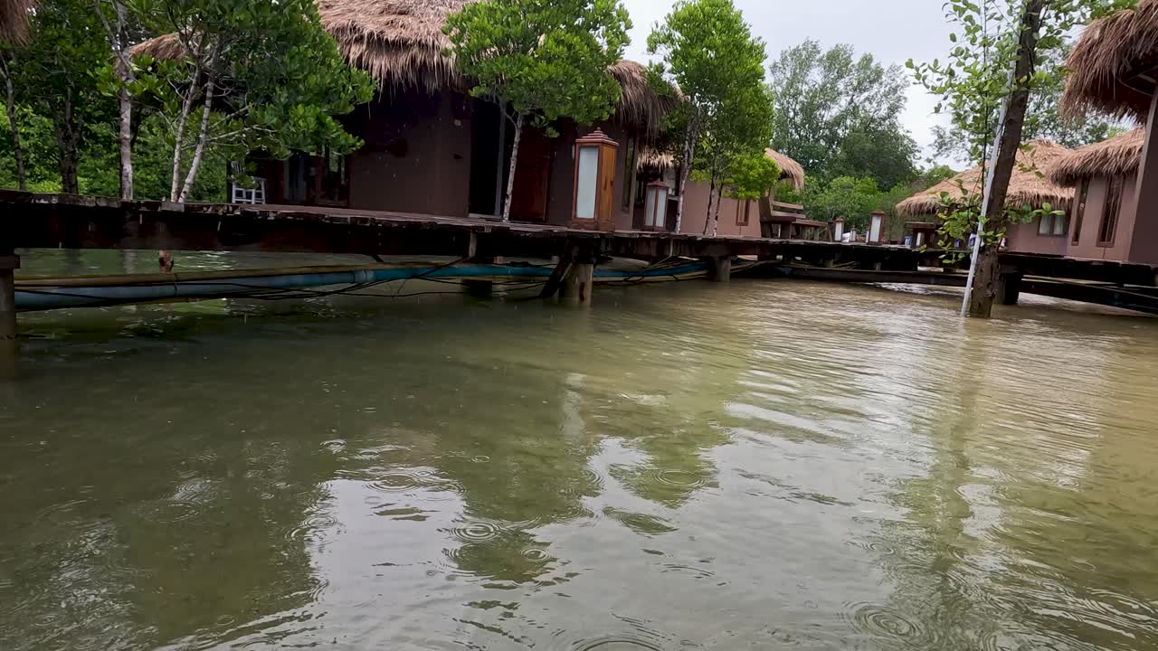 A camera smoothly transitions from above to below water near wooden bungalows and mangrove trees, revealing a calm, sandy underwater wetland in natural daylight