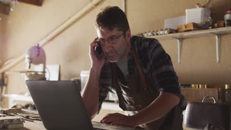 Focused-caucasian-male-knife-maker-in-workshop-using-laptop-and-smartphone