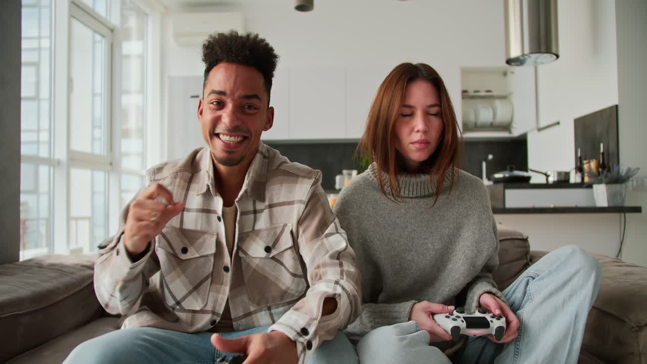 Happy and cheerful man with Black skin color a young brunette with stubble in a cream checkered shirt is very happy about his Victory in a video game while sitting with his girlfriend in a sweater on a gray sofa in a modern apartment