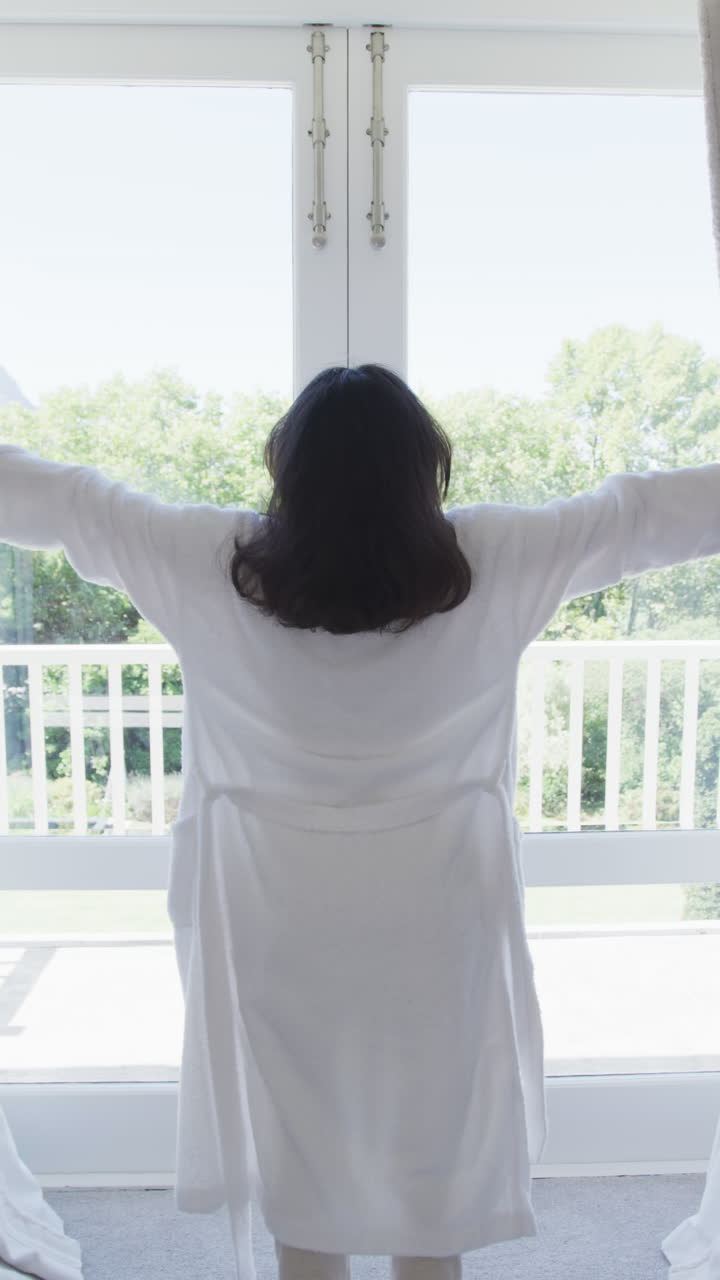 Vertical video of happy asian woman opening curtains in bedroom, in slow motion