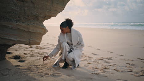 thoughtful woman drawing sand at ocean cliff. black hair traveler resting beach