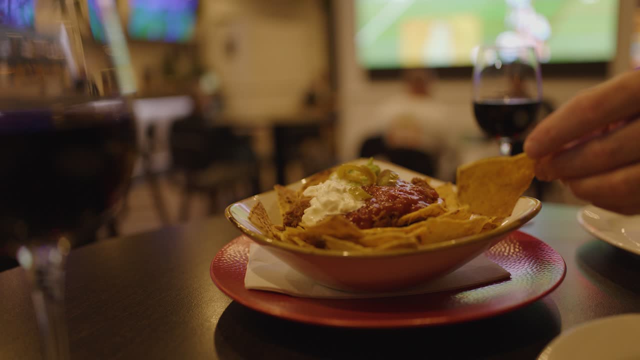 Mano che intinge una tortilla chip nella salsa nacho con vino, partita sportiva sfocata sullo sfondo della TV