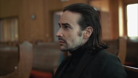 dark haired sombre man in black suit sitting in pew in church in cinematic slow motion