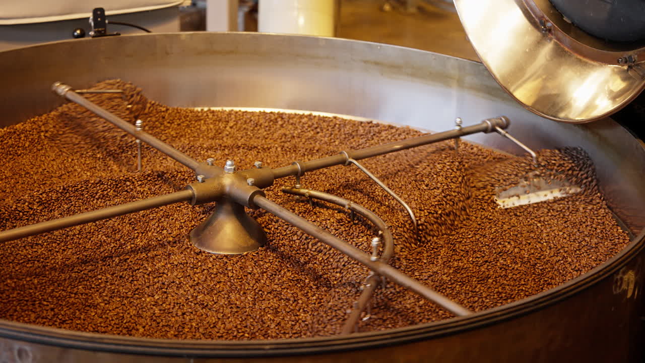 Close-up de grãos de café sendo torrados em um torrador industrial de tambor, mexidos sob calor. A textura cinematográfica quente realça o aroma fresco e a produção de café especial.