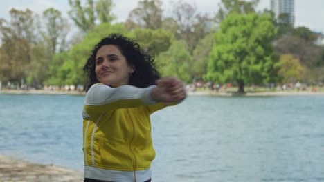 atractiva joven activa sonriendo y estirando los brazos después de un entrenamiento cerca de un lago