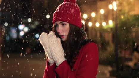 vista de cerca de una mujer caucásica con abrigo rojo y guantes calentándose las manos en la calle mientras nieva en navidad