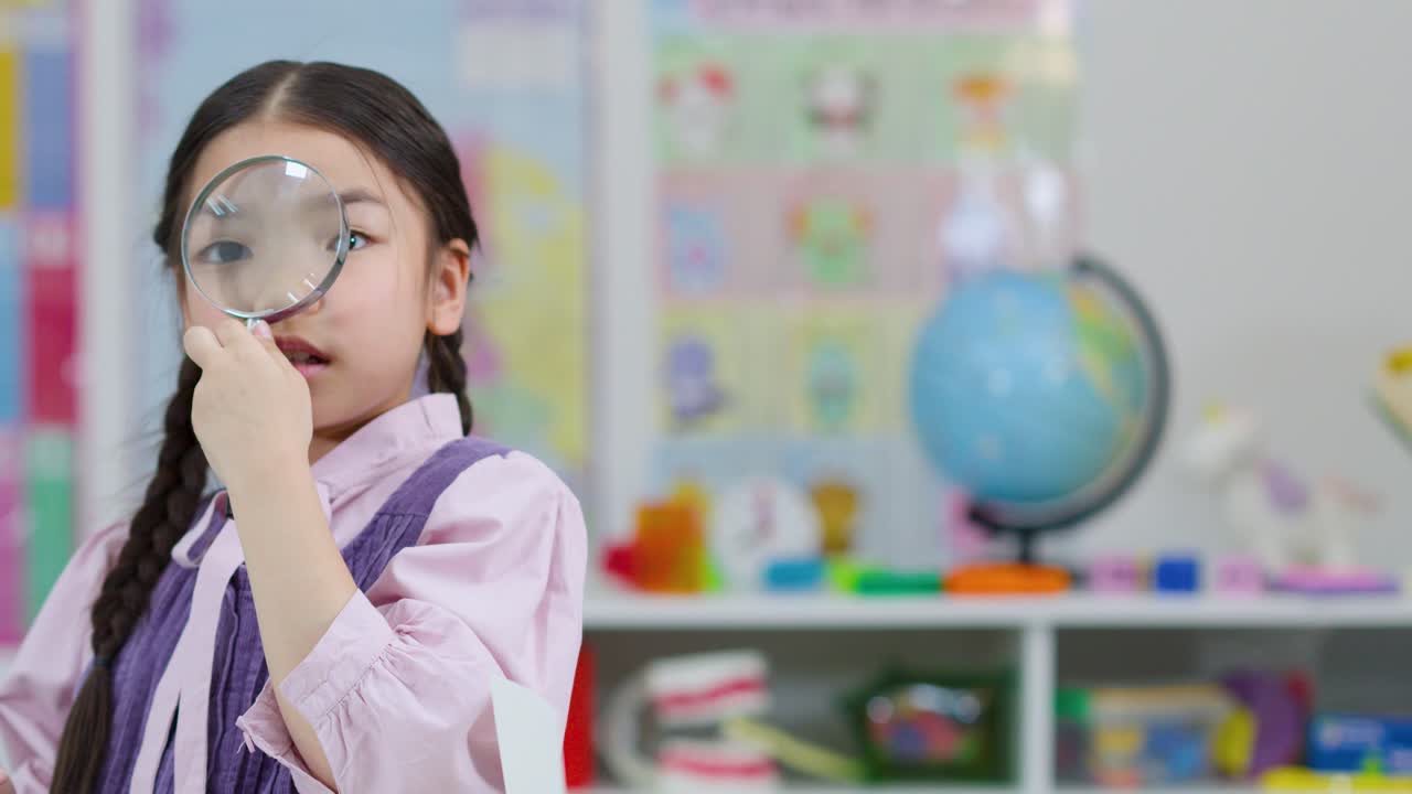 Young girl inspects through magnifying glass in colorful classroom, bright lighting, static camera