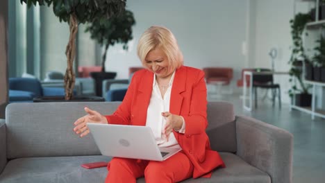 older woman in a red business suit, sitting on a couch in a business center, angrily closes her laptop, waving her hands – she's had enough