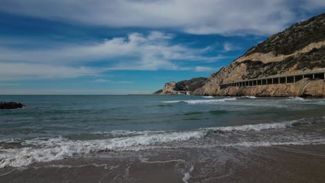 Waves-gently-lap-the-shore-by-rocky-cliffs-and-a-coastal-road-at-Port-Ginesta,-Barcelona