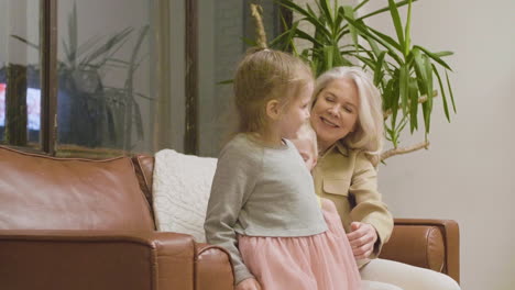 happy grandmother sitting on sofa and hugging her two little granddaughters at home