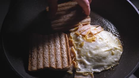 un primer plano de pan tostado que se coloca en una sartén junto con un huevo frito cubierto con jamón y queso derretido, el relleno se coloca sobre el pan para completar el ensamblaje del sándwich