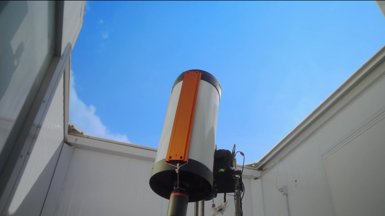 Vista dal basso dall'interno di un osservatorio con tetto scorrevole, che guarda un telescopio astronomico bianco puntato verso un cielo azzurro brillante con nuvole in rapido movimento