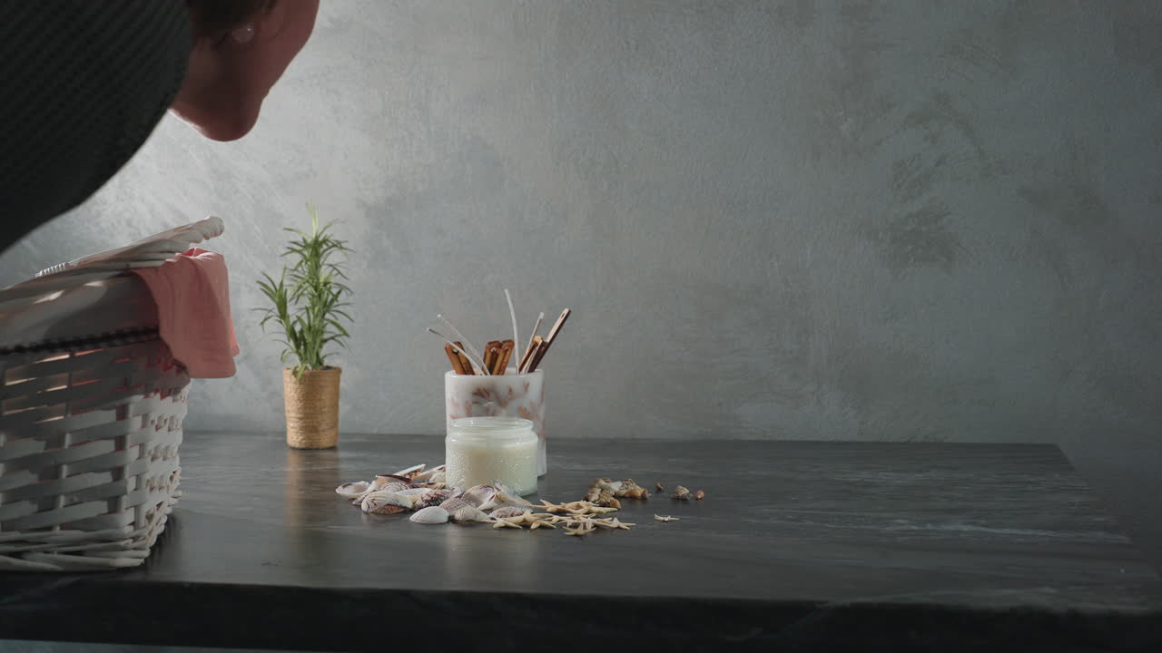 Woman blows out candle on dark marble table decorated with seashells, starfish, snail shells, and natural decor items as smoke rises