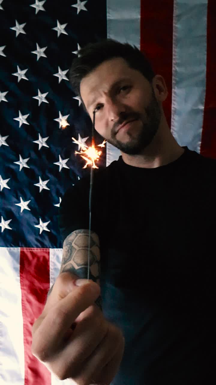 Tattooed man with a beard holding up a small sparkler that is burning to celebrate 4th of July the American birhday with a smile and happiness in front of a vertical American flag in slow motion.